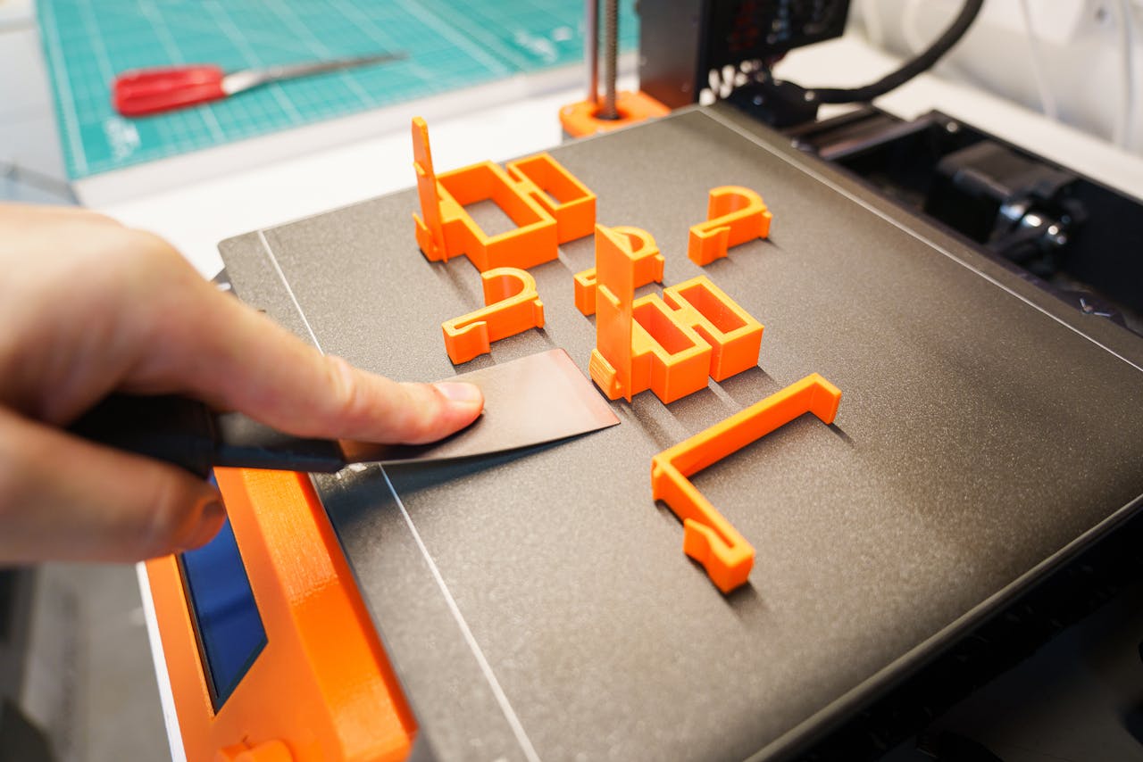 why-choose-us A hand carefully removes an orange 3D printed object from a printer bed using a spatula indoors.